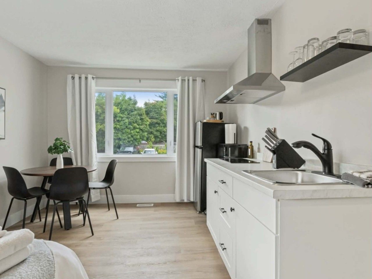 A modern kitchen with a dining area, a window with curtains, and a view of greenery outside.