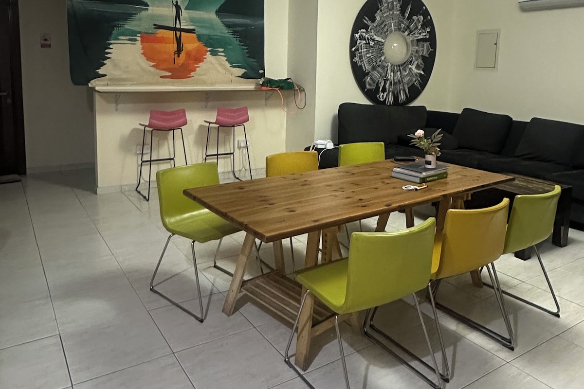 A dining area with a wooden table and chairs, a painting on the wall, and a black sofa.