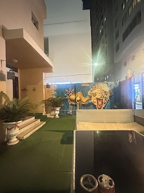 A hotel courtyard with artificial grass, potted plants, a decorative camel sculpture, and a modern building facade.