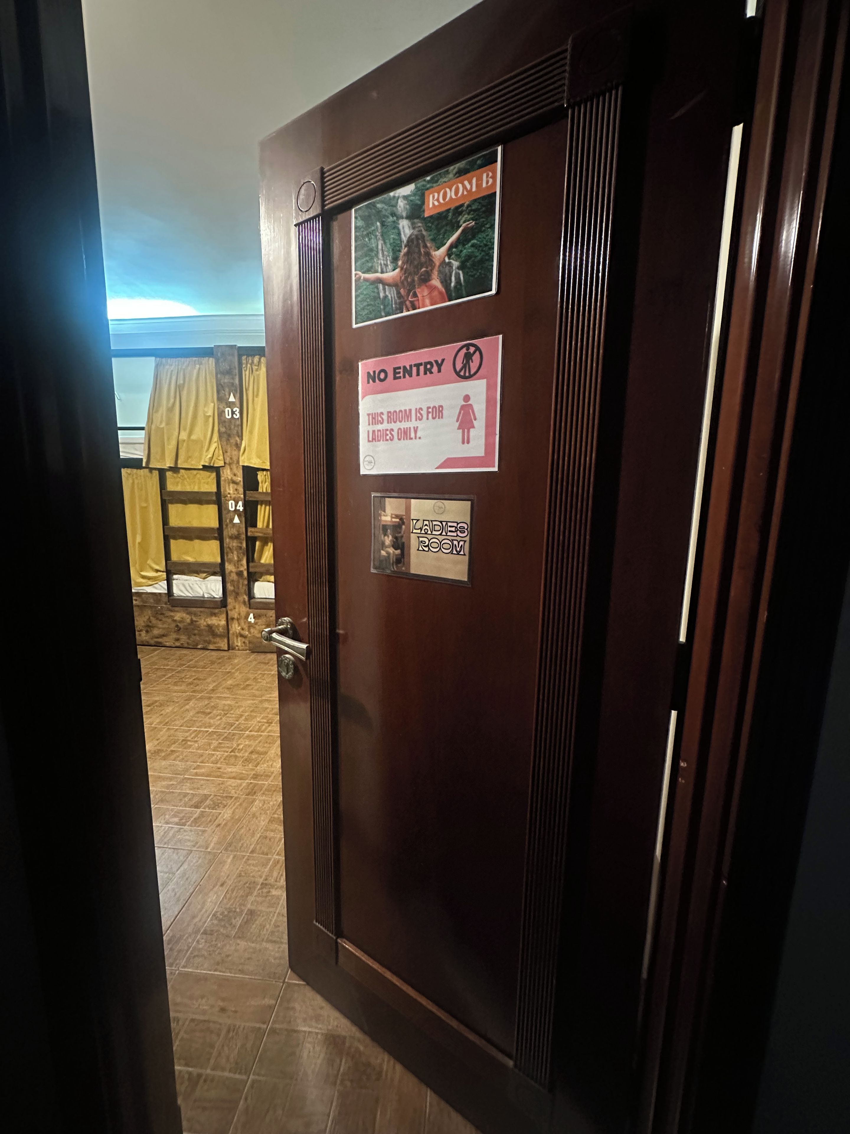 Puerta de habitación de hotel con un cartel que indica que es un baño de mujeres, un letrero de 'prohibido el acceso' y una imagen de una habitación con cama y cortinas.