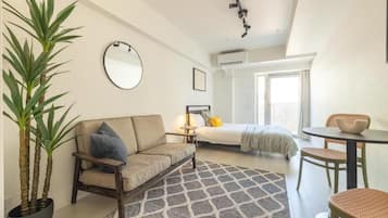 A compact hotel room with a bed, a sofa, a dining table, and a potted plant.