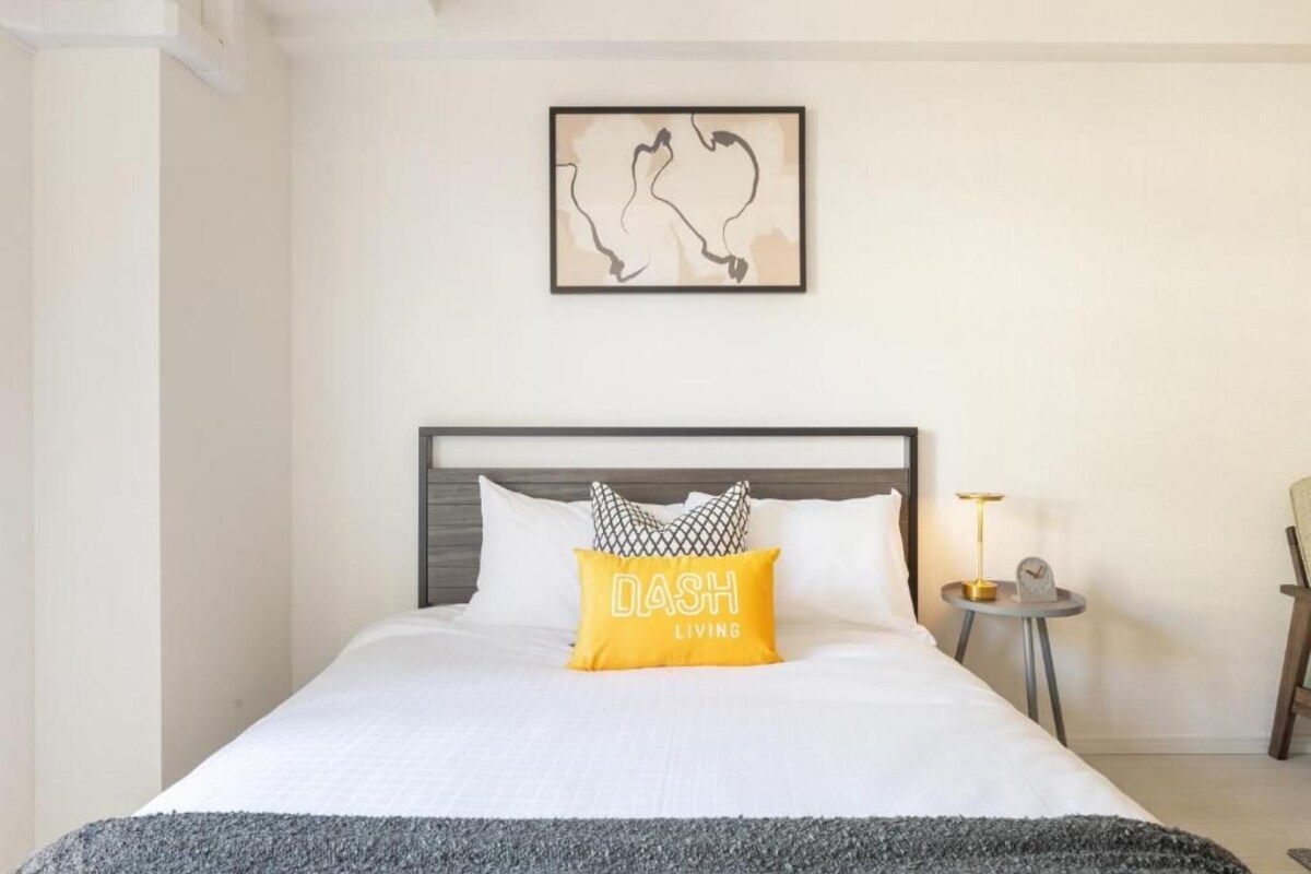 Una cama bien tendida con una colcha blanca, un plaid gris y una almohada amarilla con el texto 'DASH LIVING'.