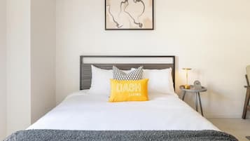 Ein ordentlich bezogenes Bett mit einer weißen Tagesdecke, einer grauen Zudecke und einem gelben Kopfkissen mit dem Aufdruck 'DASH LIVING'.