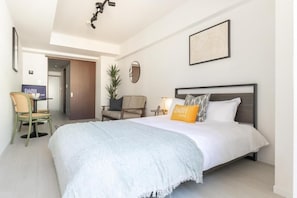 Ein Hotelzimmer mit einem Bett, einem Schreibtisch, einem Stuhl, einer Pflanze und einem gerahmten Bild an der Wand.