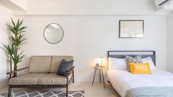 A bedroom with a bed, a sofa, a small table with a lamp, a mirror, and a potted plant.