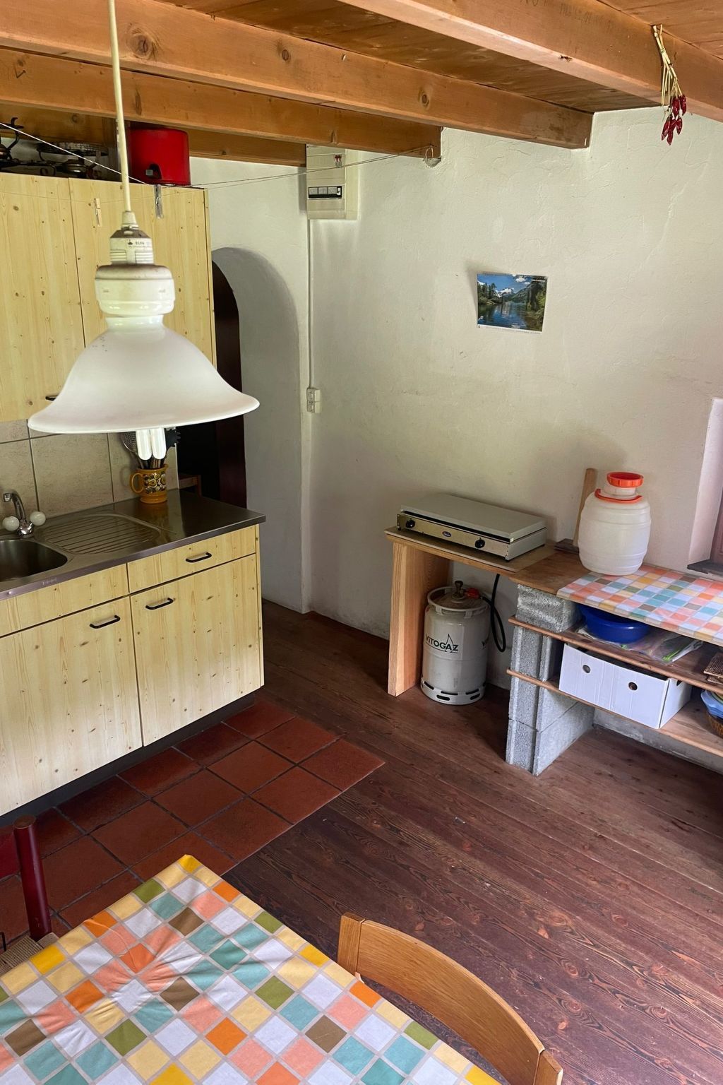 A compact kitchen with wooden cabinets, a sink, a table with a colorful tablecloth, and a gas cylinder.