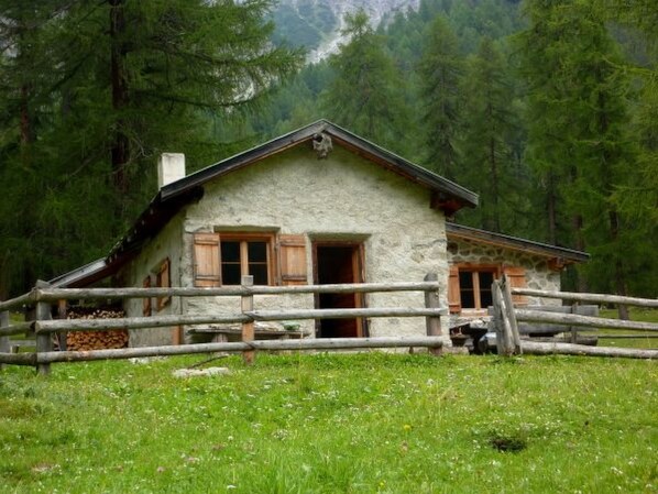 A rustic stone cabin with a wooden fence, surrounded by a forested area.