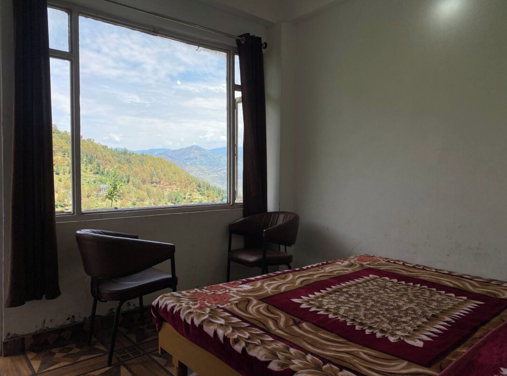 A room with a bed, two chairs, a window with a mountain view, and a patterned bedspread.