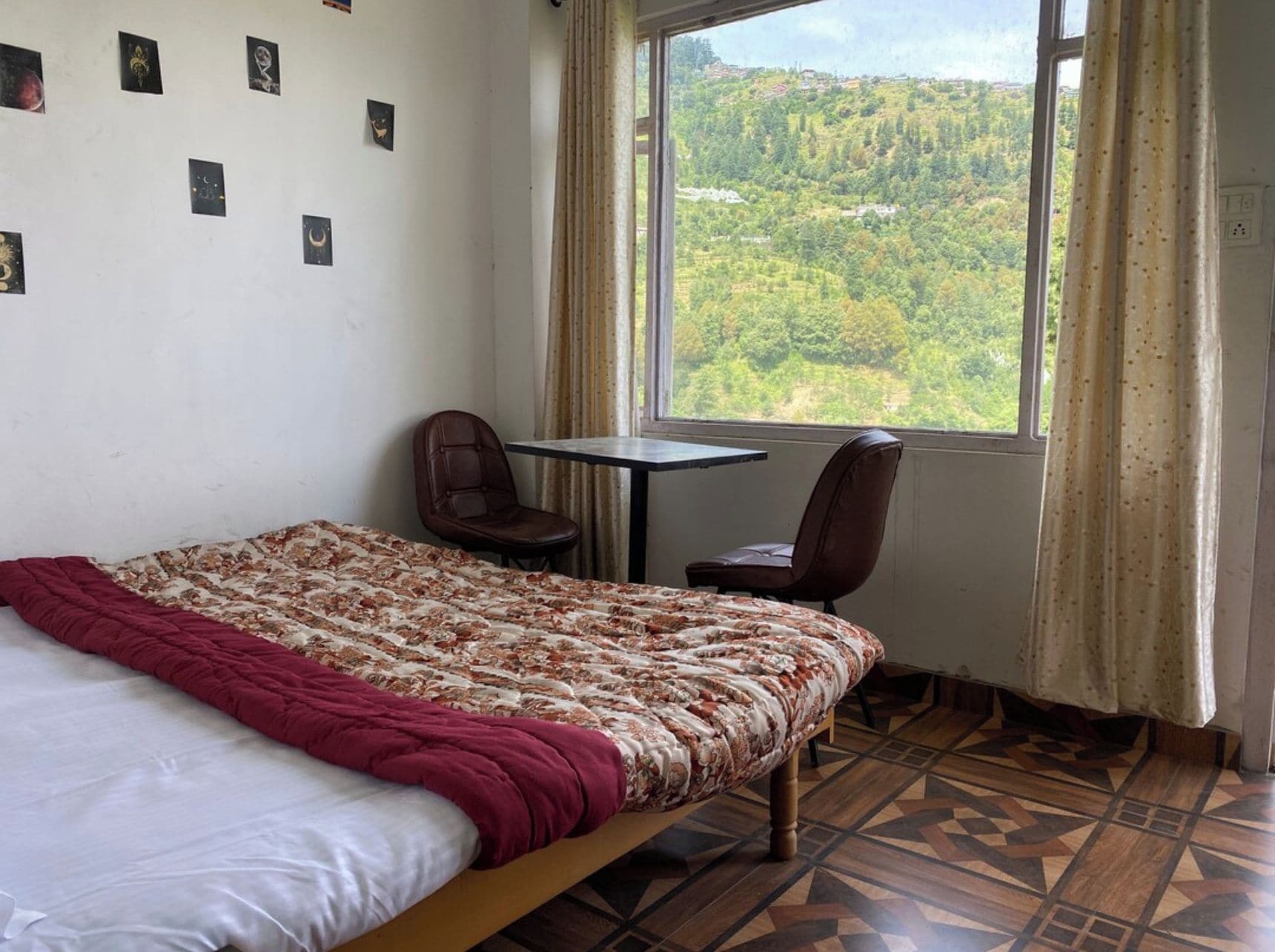 A room with a bed, a table, two chairs, a window with a view of a forest, and a wall with small pictures.