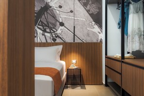 A hotel room with a bed, a small bedside table with a lamp, a wooden wardrobe, and a wall with a large abstract painting.