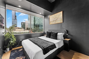 A modern bedroom with a large window, a bed, a nightstand, a lamp, and a painting. - Black Cave | Skyline Luxury 1BR • U/G Parking (Edmonton)