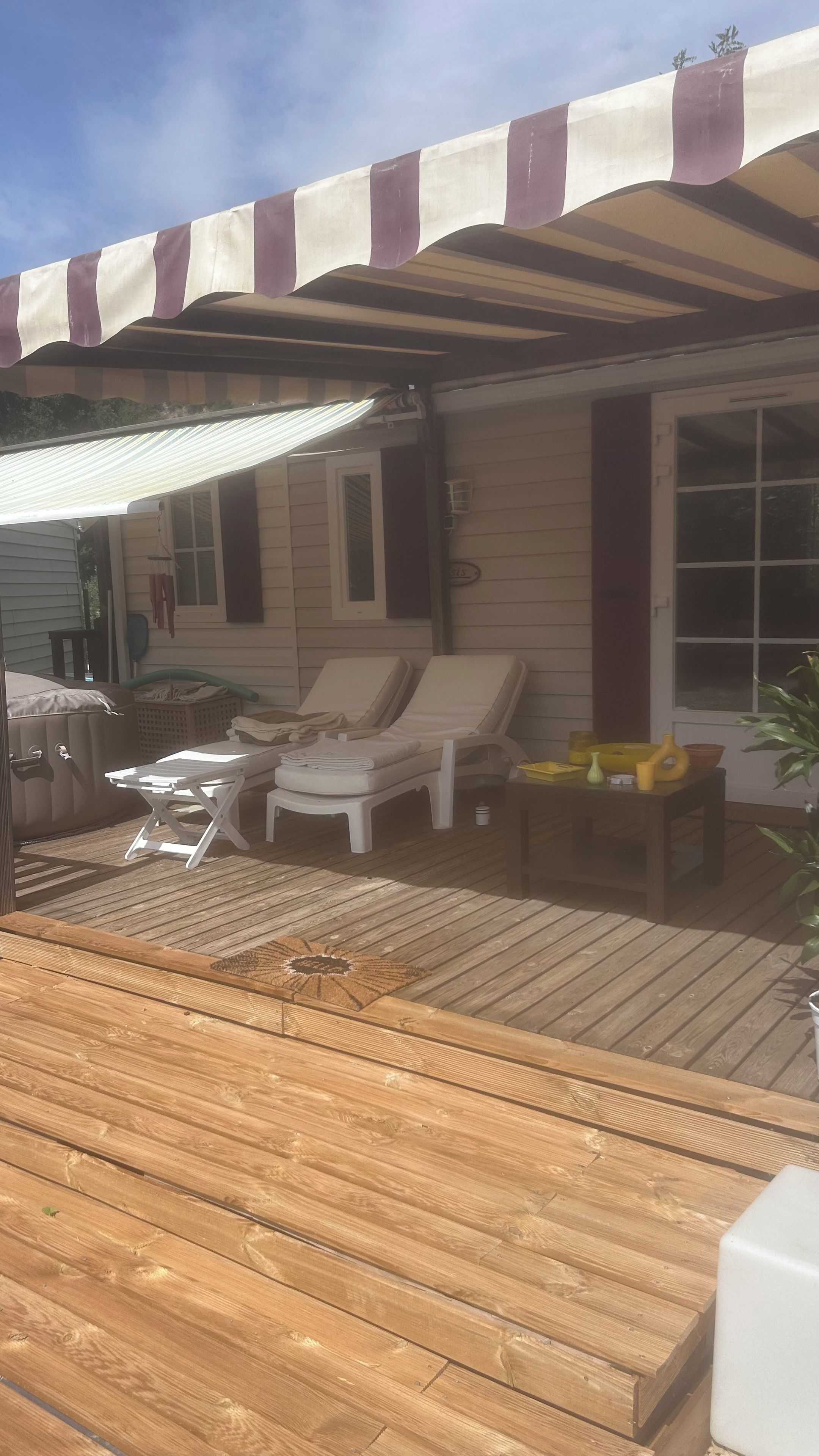 Een overdekt terras met een houten vloer, twee comfortstoelen, een kleine tafel en een opklapbaar strijkplank.