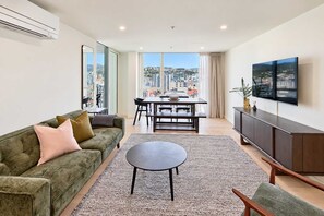 A modern living room with a sofa, a coffee table, a dining area with a table and chairs, and a TV on the wall. - Wellington CBD 3BR with Pool, Gym & Views (Wellington)