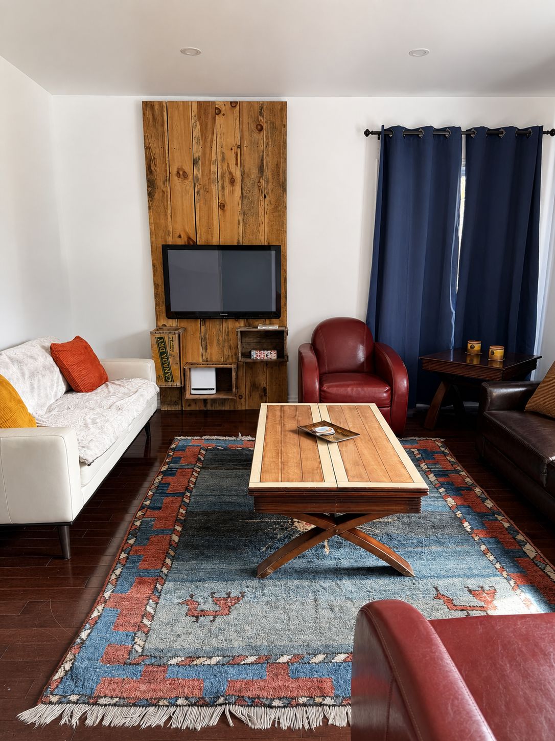 A living room with a sofa, a red chair, a coffee table, a TV mounted on a wooden wall, and a patterned rug.