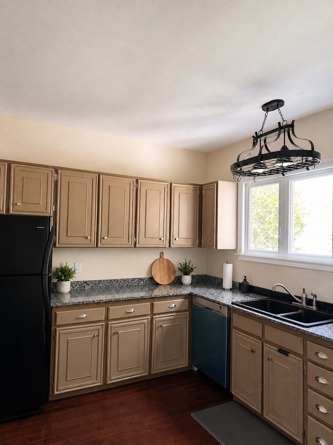 A kitchen with beige cabinets, a black refrigerator, a marble countertop, a sink, and a window with a view of greenery.