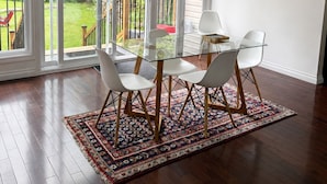 A dining area with a glass table and chairs, a patterned rug, and a view of a wooden deck and greenery outside. - Charming Chelsea Bungalow Central Location Modern Comforts (Chelsea)
