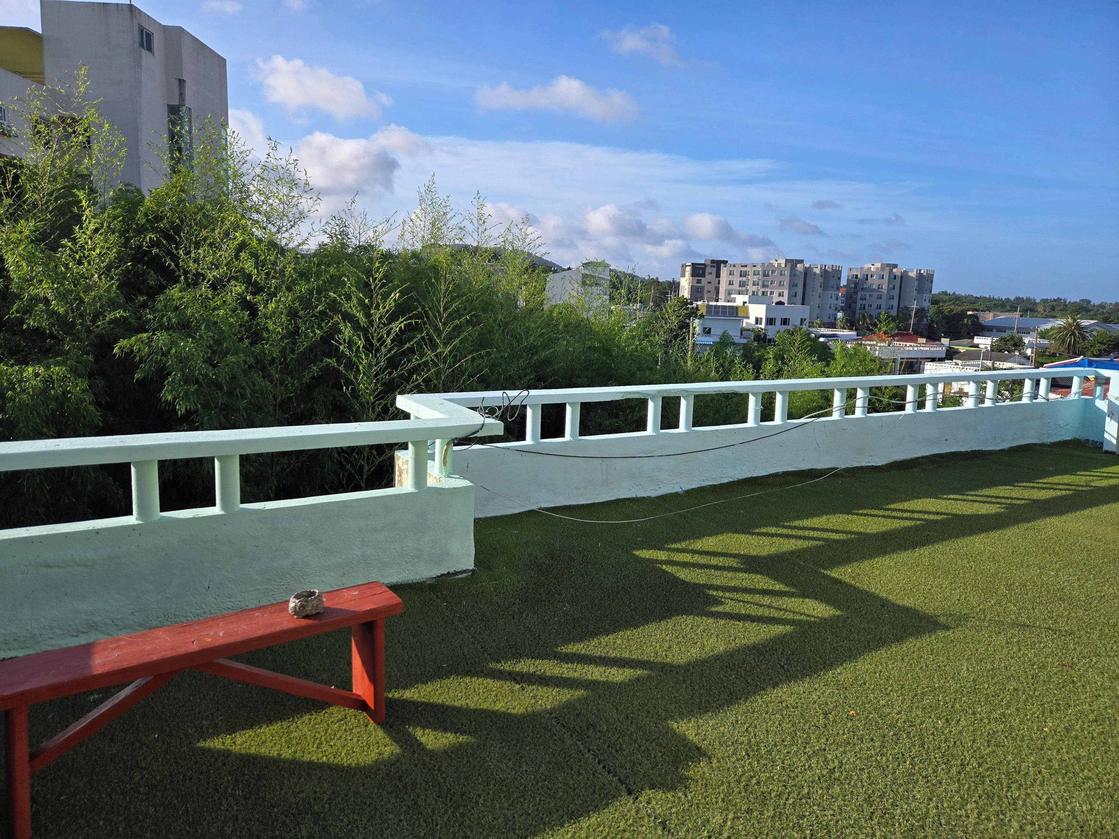 A grassy area with a white fence, a red bench, and buildings in the background.