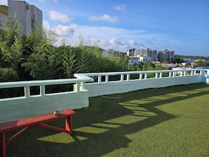 Un área verde con una valla blanca, un banco rojo y edificios al fondo.