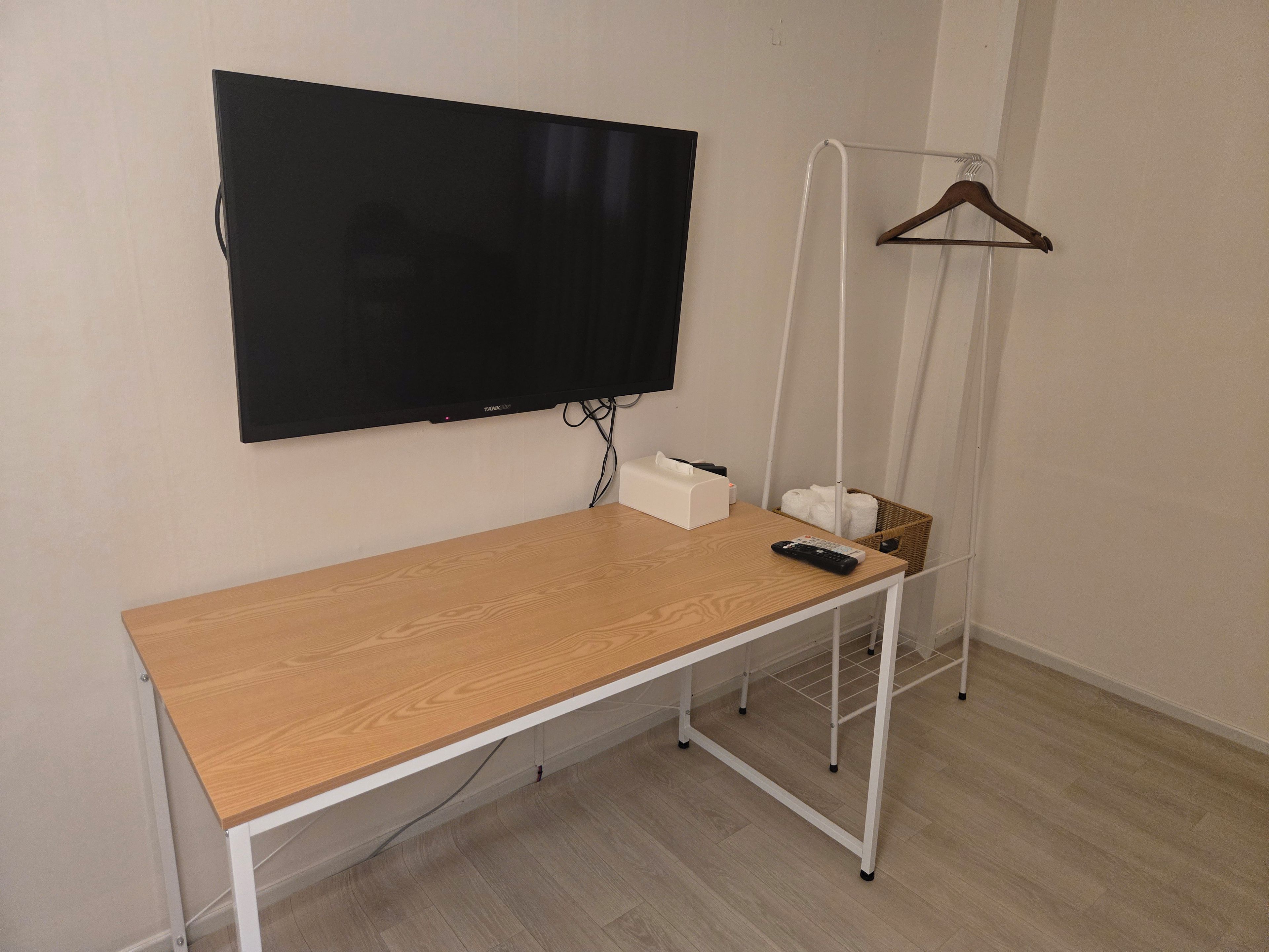 Una habitación con un escritorio de madera, un televisor montado, un perchero y una cesta con toallas.