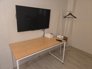 Ett rum med ett skrivbord i trä, en väggmonterad tv, en klädstång och en korg med handdukar.