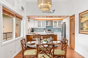 A modern kitchen with white cabinets, a central island, and a dining area with wicker chairs. - Ocean-view 1BR Kapalua condo with balcony, pool, AC, and full kitchen-Kapalua Ridge Villas 2521 (Lahaina)