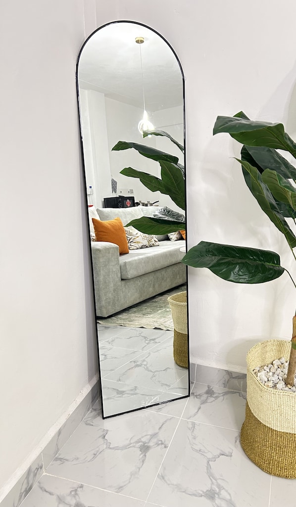 A room with a large mirror reflecting a living area, a grey sofa with cushions, a hanging plant, and a woven basket with stones.