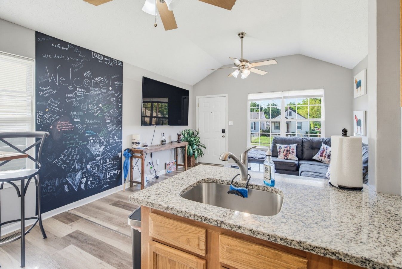 A modern kitchen with a granite countertop, stainless steel sink, and wooden cabinets. There is a chalkboard wall with various writings and a ceiling fan.