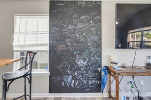 A room with a large chalkboard wall covered in handwritten messages and drawings, a window with blinds, a chair, a wooden table with various items, and a ceiling fan. - Derby House - Louisville (Louisville)
