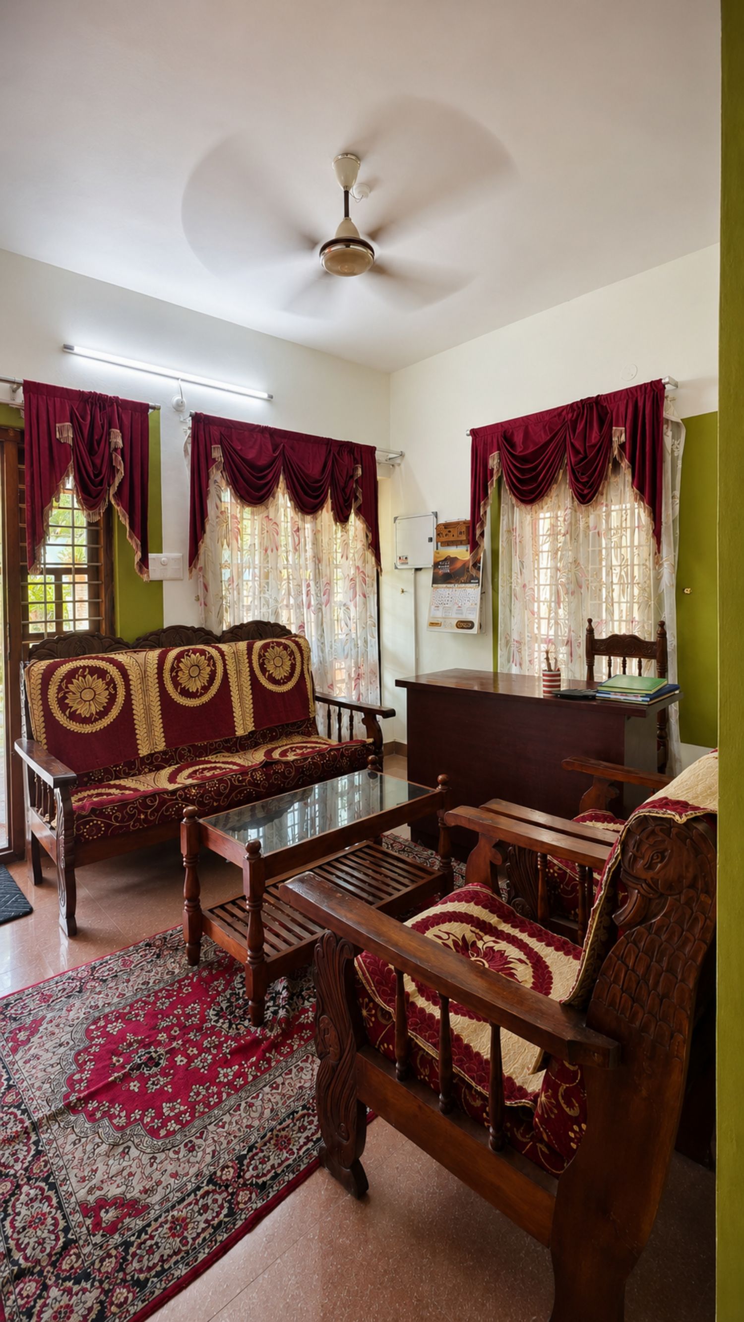 Un salón tradicional con un sofá, una mesa de madera y una silla. Hay un ventilador de techo, una ventana con cortinas y una pared verde.