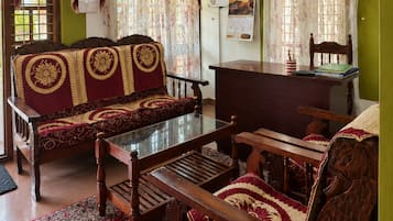 Un salón tradicional con un sofá, una mesa de madera y una silla. Hay un ventilador de techo, una ventana con cortinas y una pared verde.
