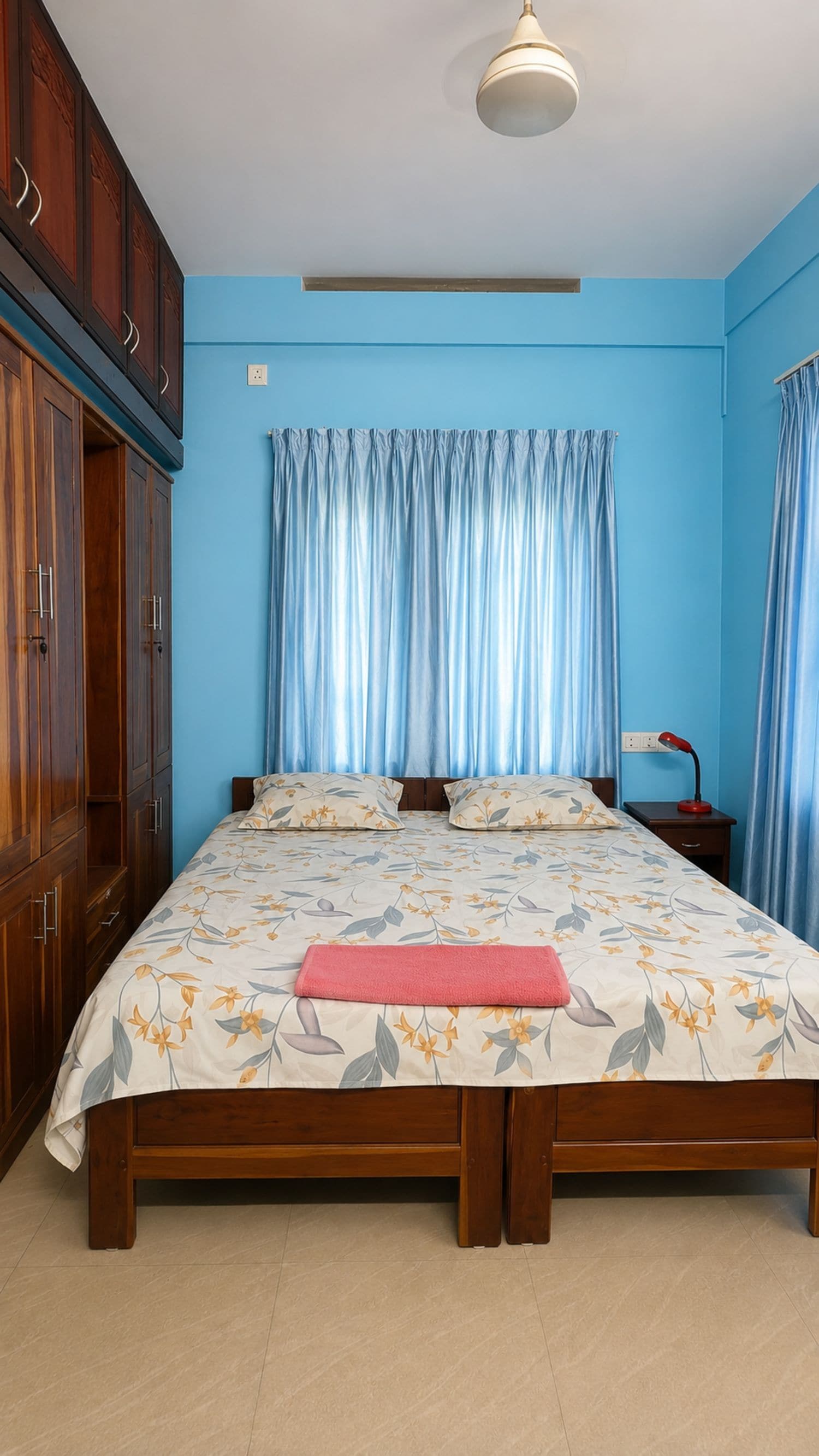 A bedroom with a bed, wooden wardrobe, curtains, and a bedside lamp.