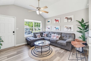 A modern living room with a sectional sofa, a glass coffee table, and horse-themed artwork on the wall. - Derby House (Louisville)