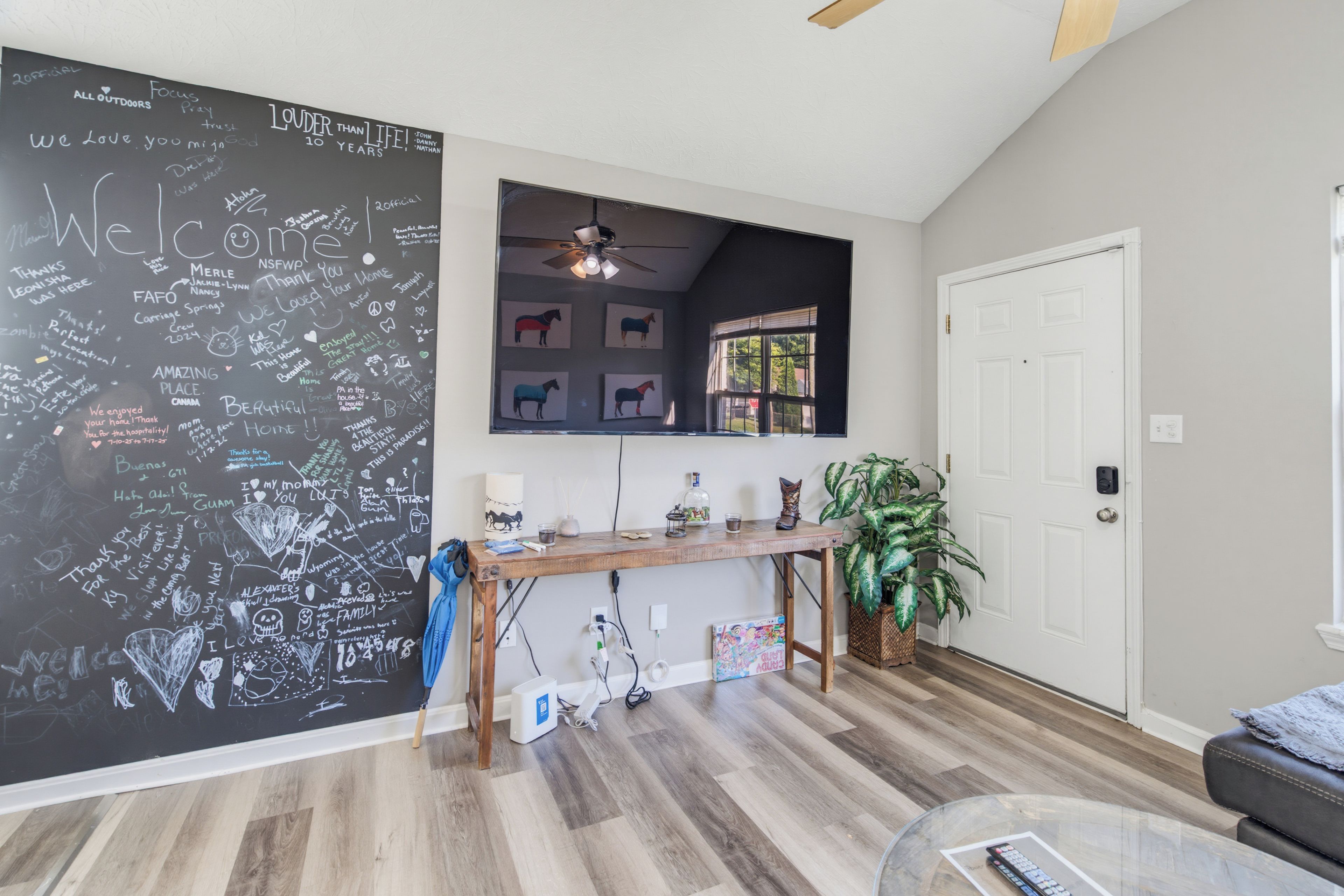 Una habitación con un muro de pizarra lleno de mensajes, una consola de madera con varios objetos, un ventilador de techo y una puerta con ventana.