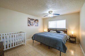 Ein Schlafzimmer mit einem Babybett, einem Bett, einem Deckenventilator, einem Fenster mit Jalousien und einem Wandbild.