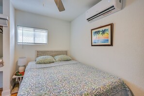 Ein Schlafzimmer mit einem Bett, einem Fenster mit Jalousien, einer Klimaanlage, einer Nachttischlampe und einem gerahmten Bild an der Wand.
