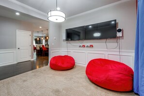 A modern living room with a flat-screen TV mounted on the wall, two red bean bags, and a dining area in the background. - McDonough Group Getaway: Pool, Patio + Game Room (McDonough)