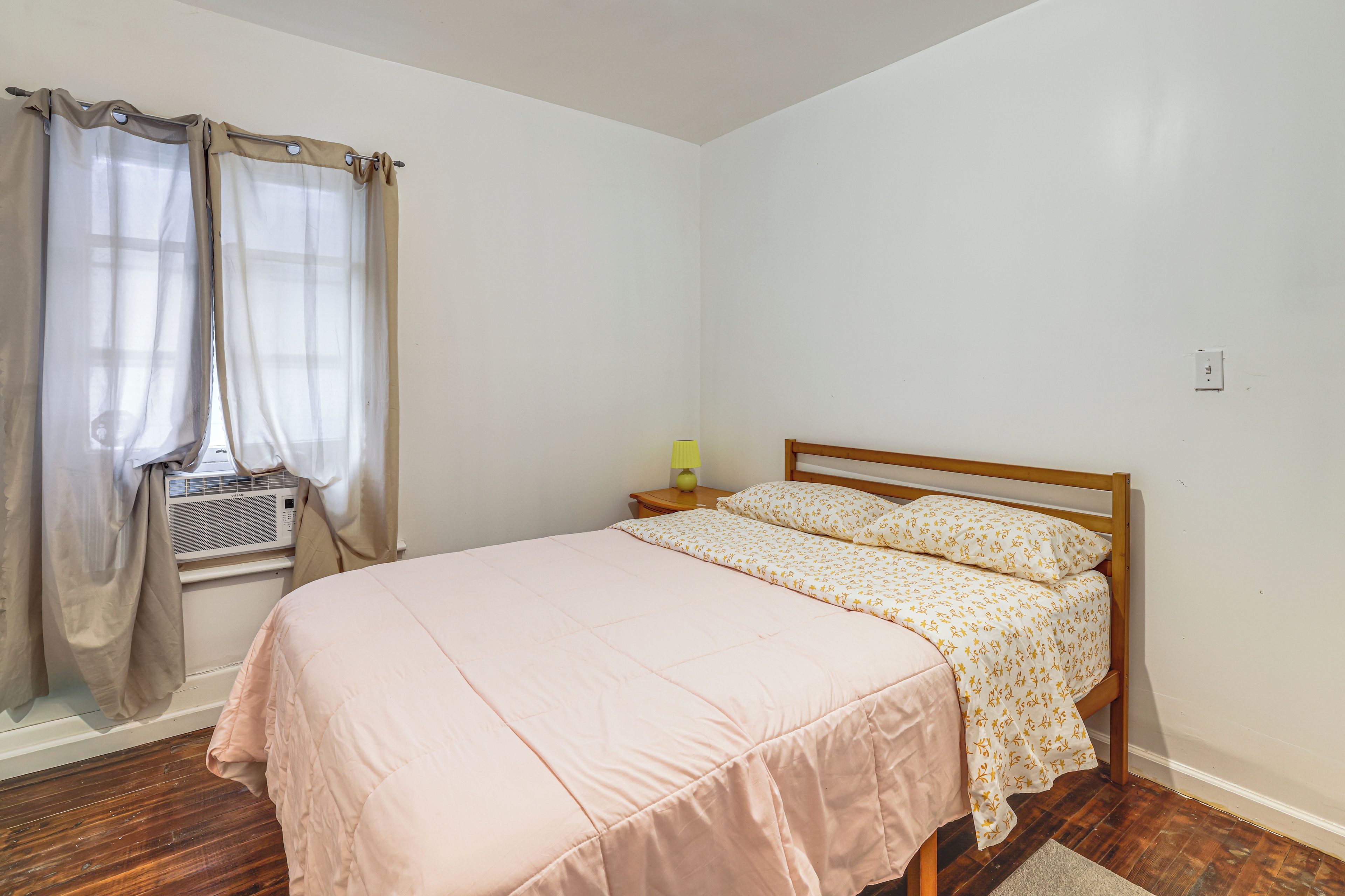 A bedroom with a bed, a nightstand, a lamp, and an air conditioning unit.