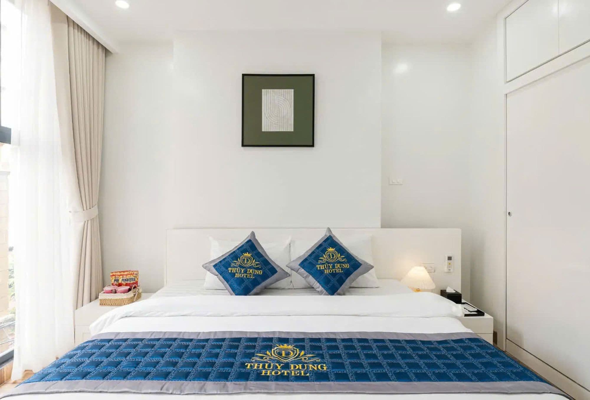 Quarto de hotel com cama, almofadas, mesa de cabeceira, luminária e quadro pendurado na parede.