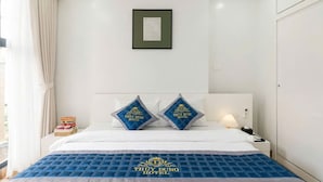 Quarto de hotel com cama, duas almofadas com o logotipo do hotel, uma moldura na parede e uma mesa de cabeceira com abajur.