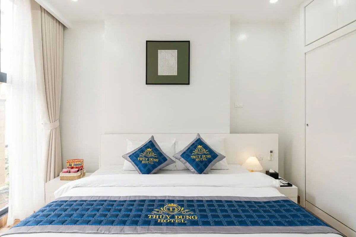 A hotel room with a bed, two pillows with the hotel's logo, a framed picture on the wall, and a nightstand with a lamp.