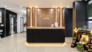 Balcão de recepção de hotel com design moderno, placa digital exibindo o nome do hotel e um pequeno santuário com oferendas.