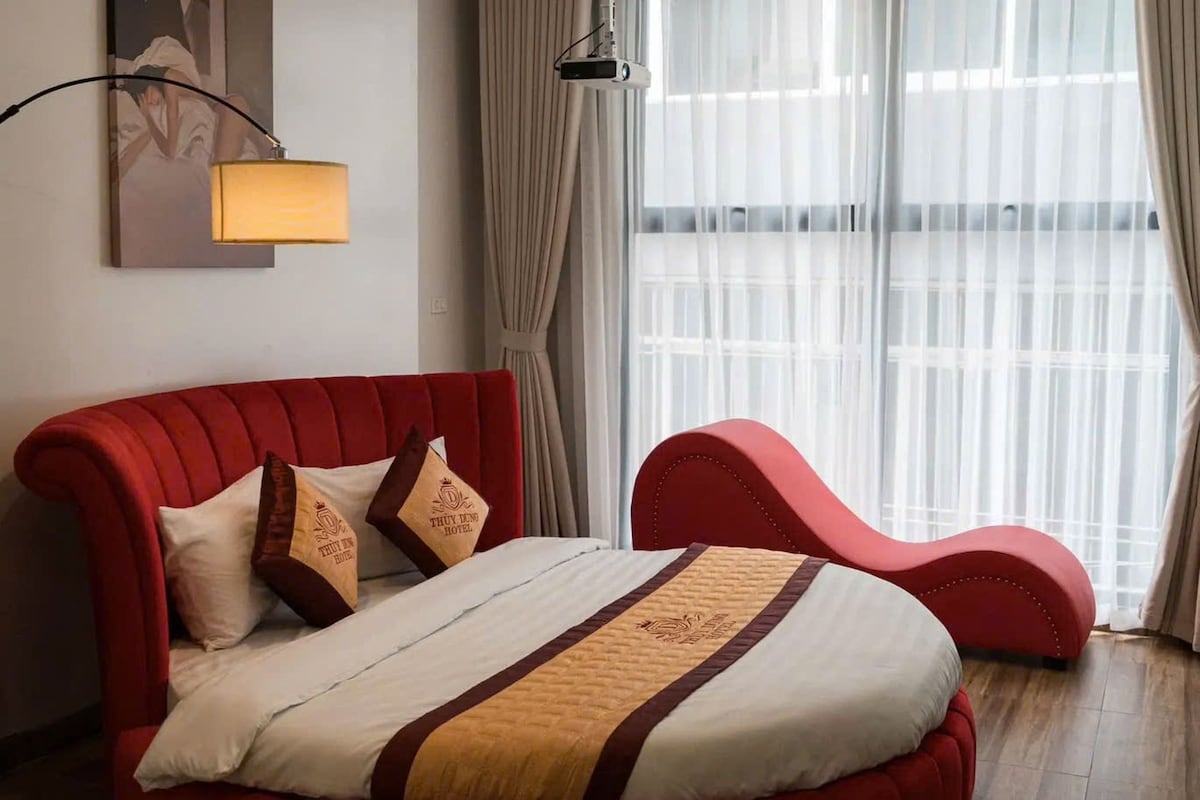 A bedroom with a red upholstered bed, a bedside lamp, and a large window with curtains.