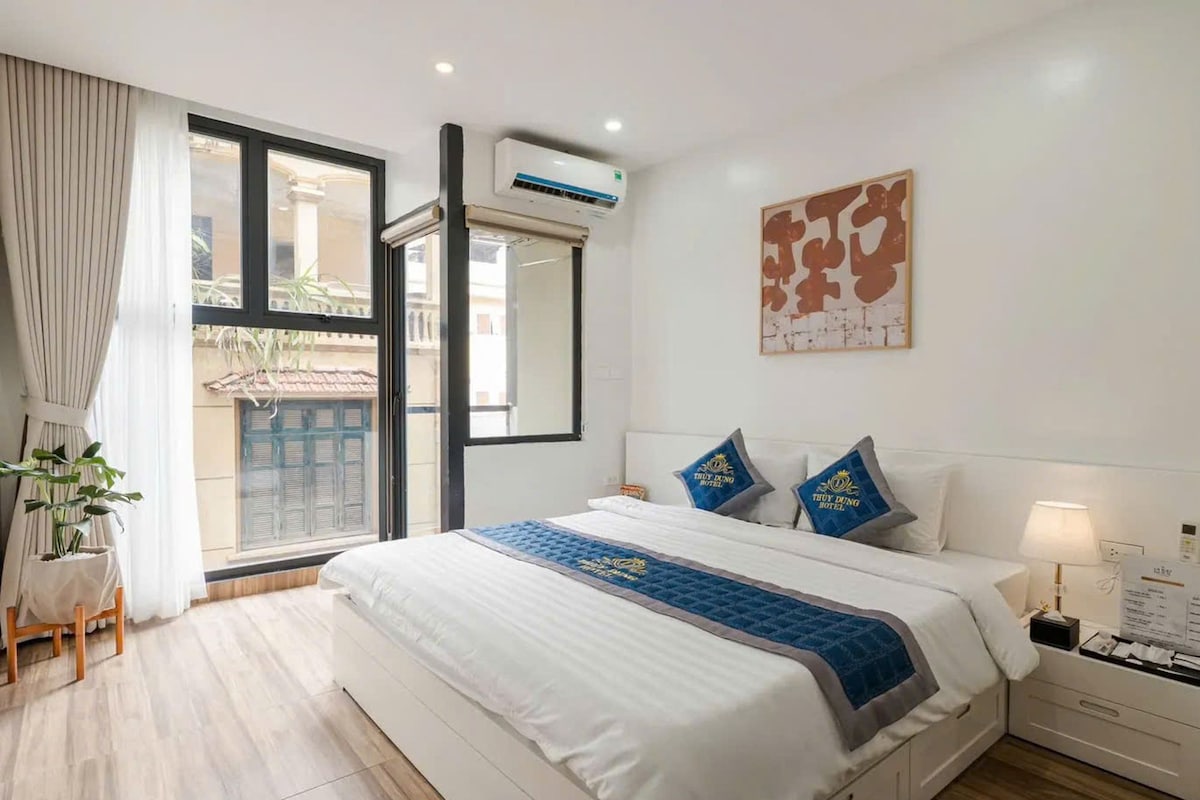 A hotel room with a bed, a nightstand, a lamp, a window with curtains, and a wall-mounted air conditioner.