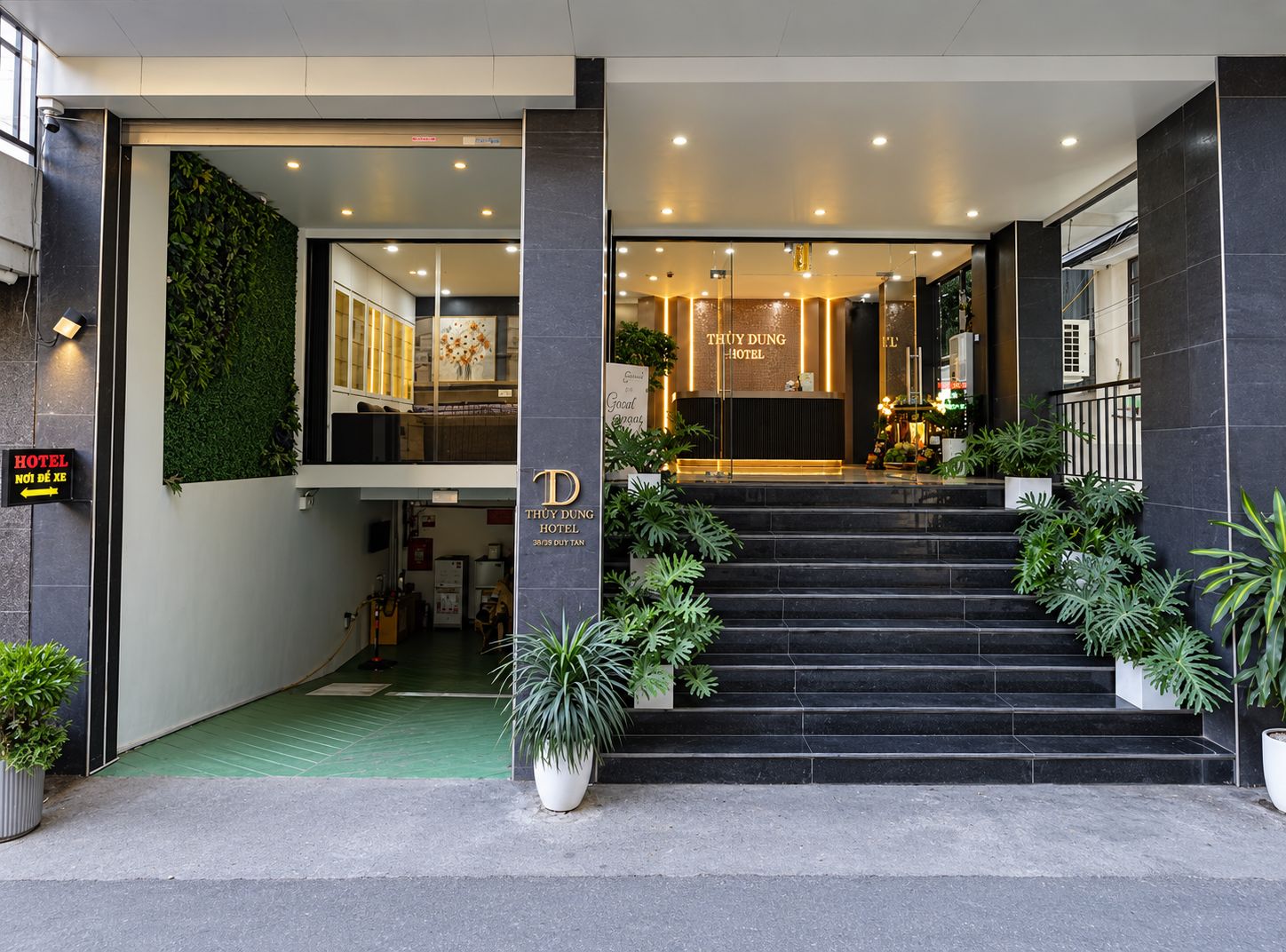 Entrada de hotel com balcão de recepção, escada que leva ao lobby e plantas em vasos.