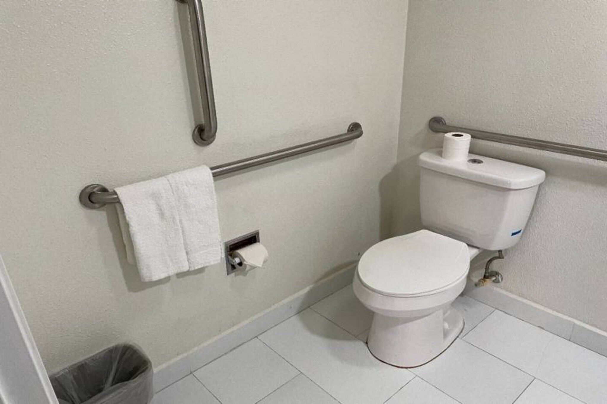 A standard bathroom with a toilet, handrails, a towel rack, and a trash bin.