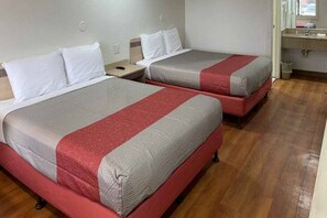 Duas camas com roupa de cama vermelha e cinza, um piso de madeira e uma pequena mesa de cabeceira com abajur.