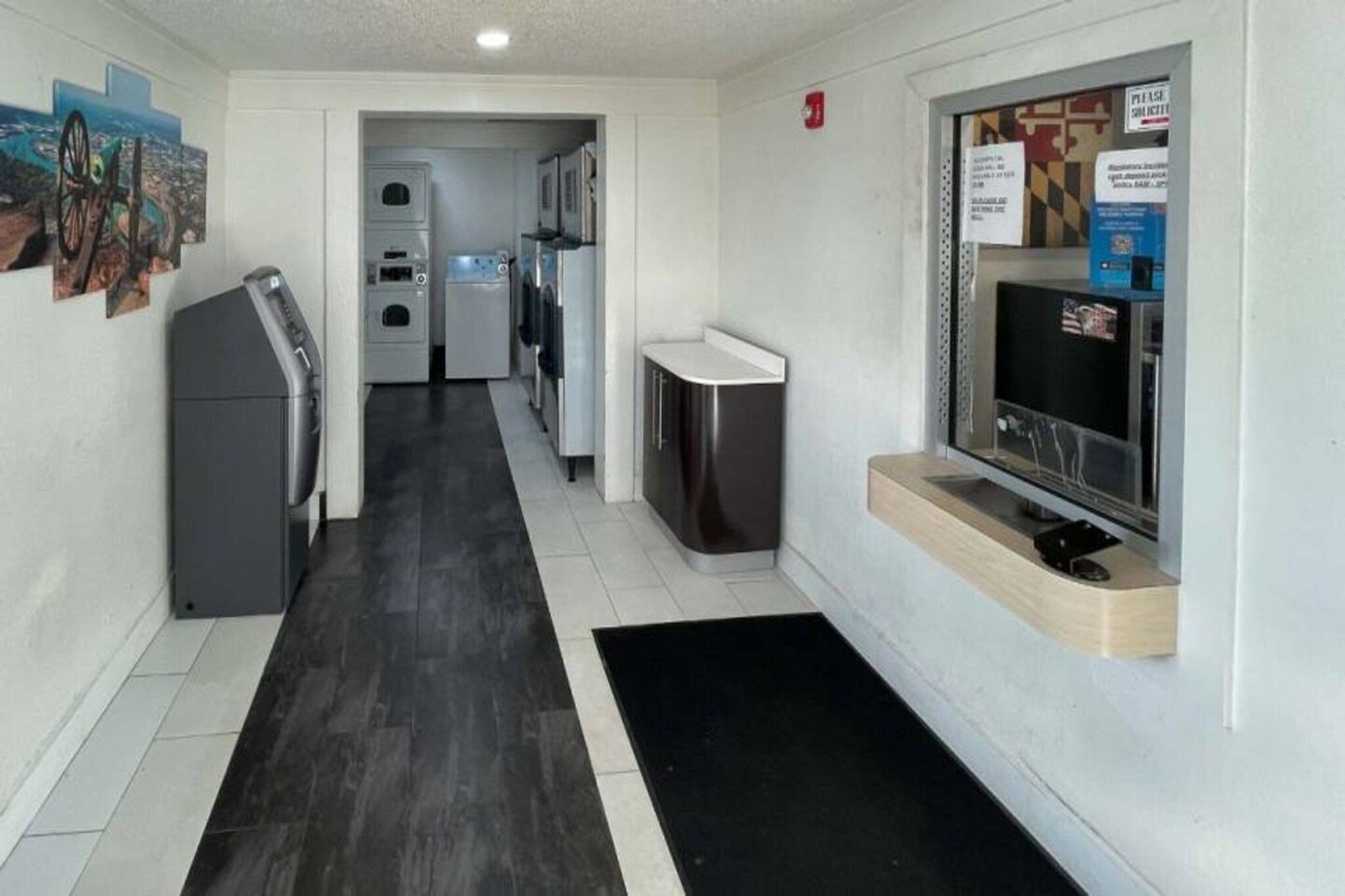 A hallway with a black mat leading to a kitchenette area, a refrigerator, and a television on a wooden shelf.