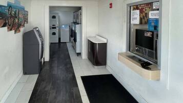 A hallway with a black mat leading to a kitchenette area, a refrigerator, and a television on a wooden shelf.