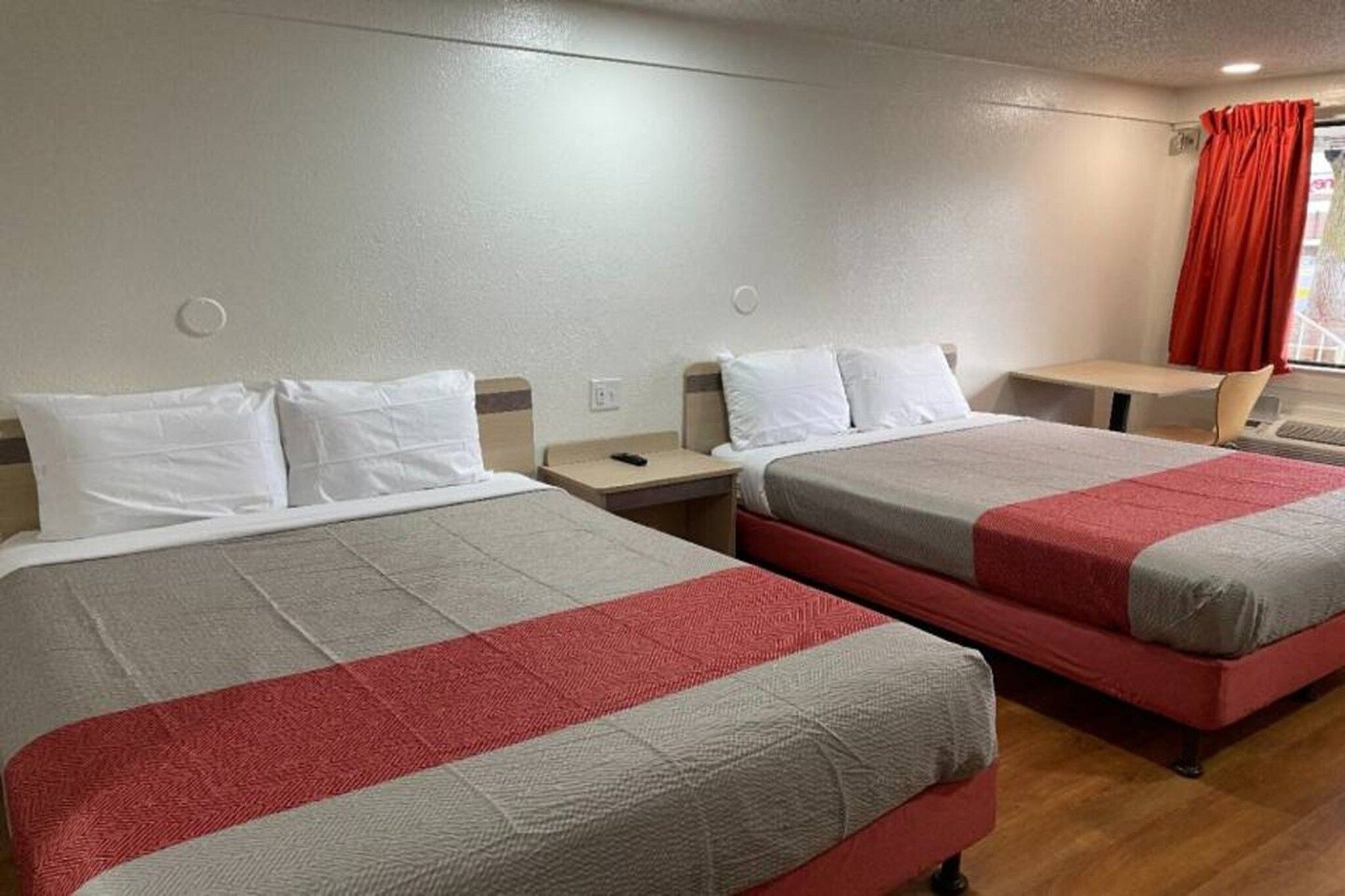 Et hotelværelse med to senge, hver med hvide puder og sengetøj i røde og grå farver. Der er et lille bord og en stol i hjørnet.