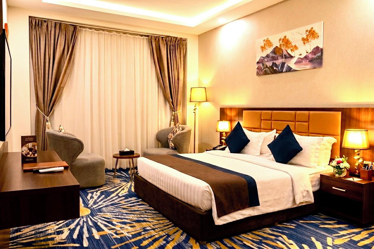 Una habitación de hotel con una cama tamaño king, dos sillones, un escritorio, una lámpara y un cuadro en la pared.